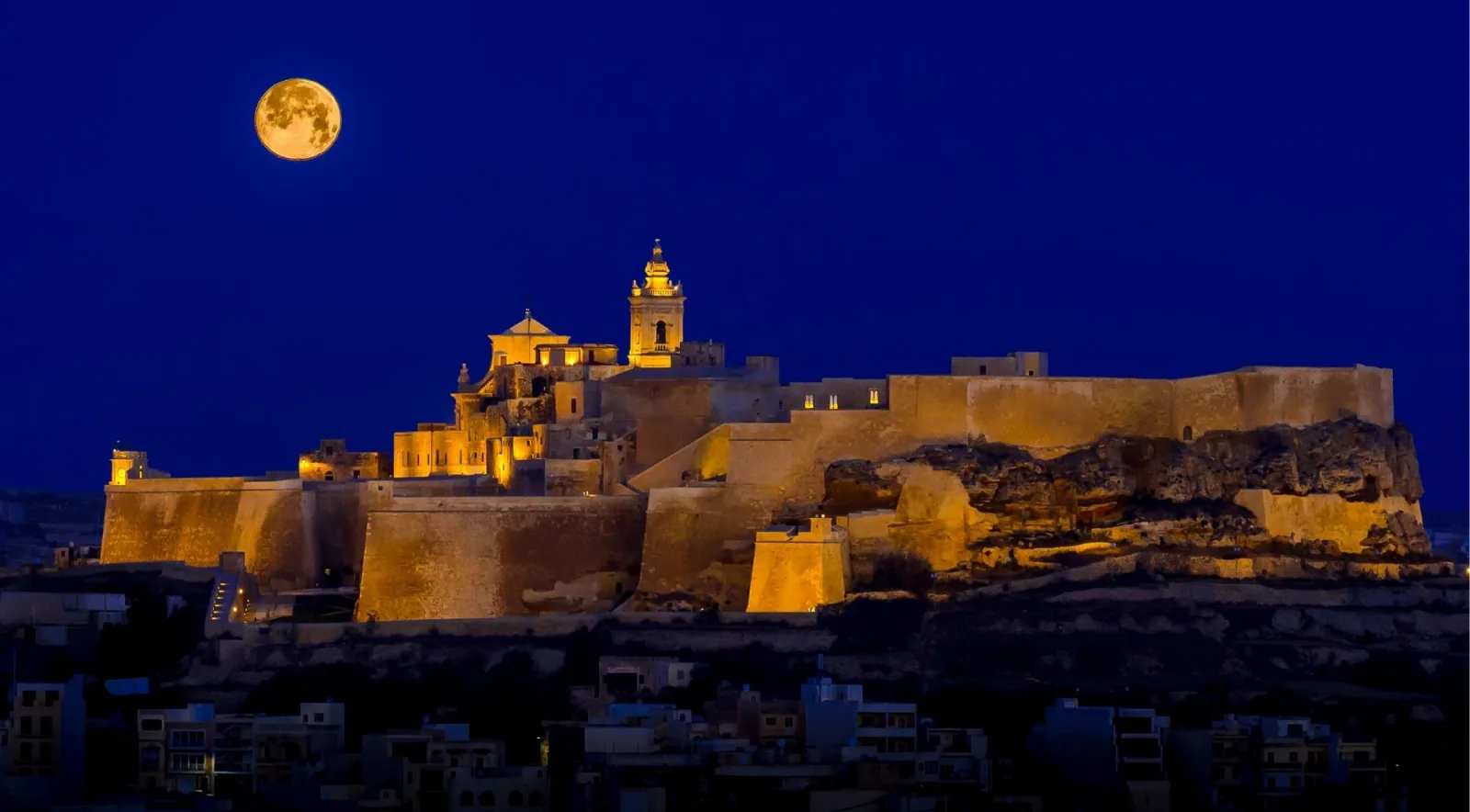 The floodlit Gozo Citadel at night beneath a full moon above Victoria