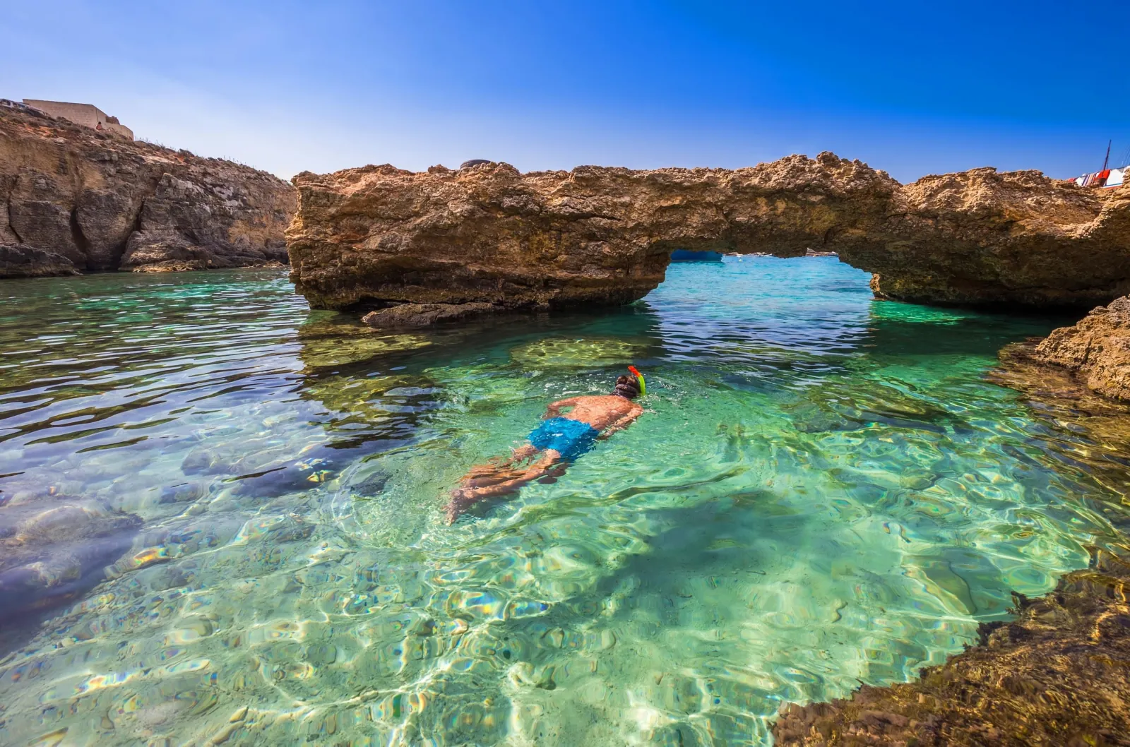 Snorkeller in clear water beneath a natural rock arch near Comino